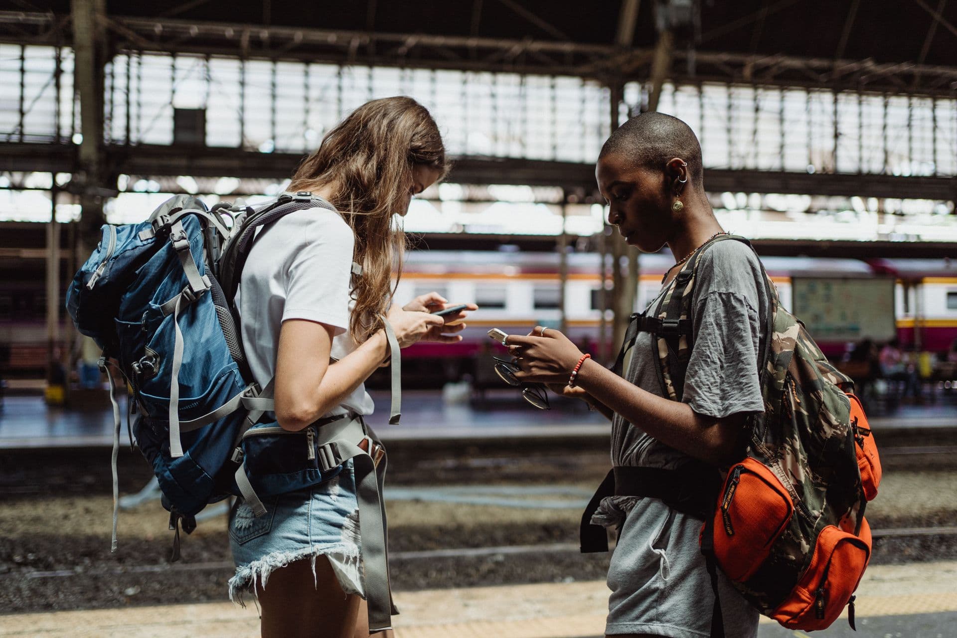 Duas intercambistas de mochilão usando os celulares em estação de trem